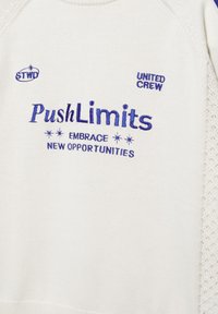 Bela pletena tkanina z modrim vezenim besedilom: "STWO," "UNITED CREW," "PushLimits" in "SPREJMI NOVE PRILOŽNOSTI" z zvezdastimi simboli.