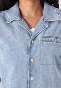 Camisa de denim azul claro com colarinho, apresentando botões prateados e um único bolso no peito. O tecido tem um acabamento ligeiramente texturizado.