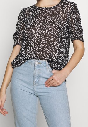 Frau trägt eine schwarze, transparente Bluse mit weißem Blumenmuster und hellblaue High-Waist-Jeans, Hand ruht in der vorderen Tasche.