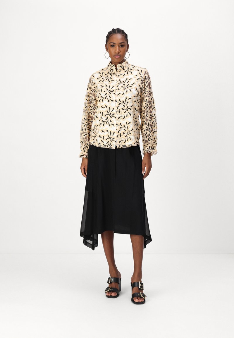 Bež jakna s vezom i cvjetnim uzorkom, dugim rukavima i nabranim rukavima, u kombinaciji s crnom asimetričnom suknjom i crnim sandalama.