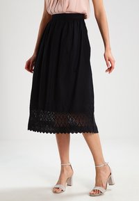 Jupe midi noire avec un ourlet en dentelle texturée, taille froncée et tissu léger. Associée à des chaussures à bout ouvert et un haut léger.