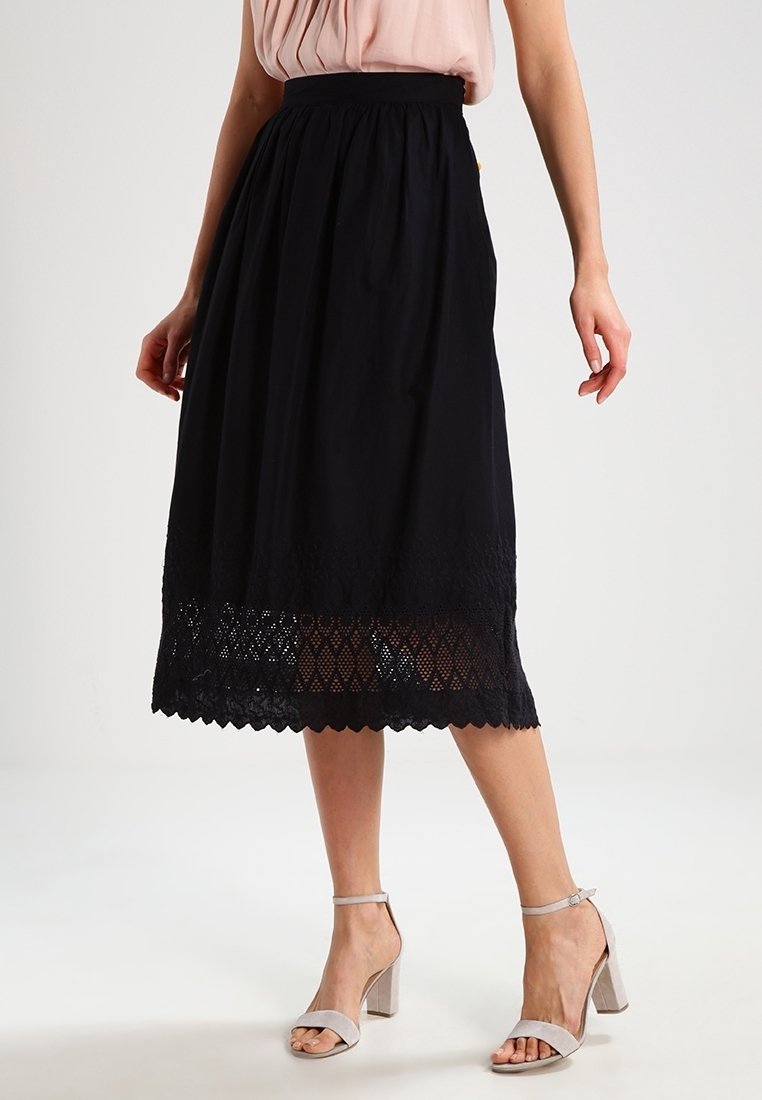 Jupe midi noire avec un ourlet en dentelle texturée, taille froncée et tissu léger. Associée à des chaussures à bout ouvert et un haut léger.
