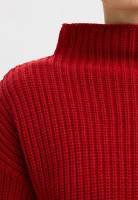 Suéter rojo de punto con cuello alto y textura acanalada, que destaca un hilo grueso y un diseño estructurado.