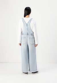 Salopette en denim bleu clair avec une coupe ample, des bretelles ajustables et deux poches arrière. Associée à une chemise blanche à manches longues et des baskets noires.
