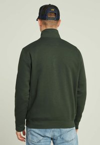 Sudadera verde oscura con cuello alto, puños y dobladillo acanalados, que presenta un tejido texturizado y una pequeña etiqueta de logo en el lado.
