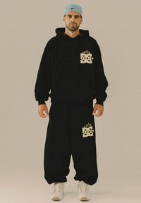 Hombre con sudadera negra y pantalones anchos negros a juego con logotipos, zapatillas blancas y gorra ligera azul al revés, de pie frente a un fondo uniforme.