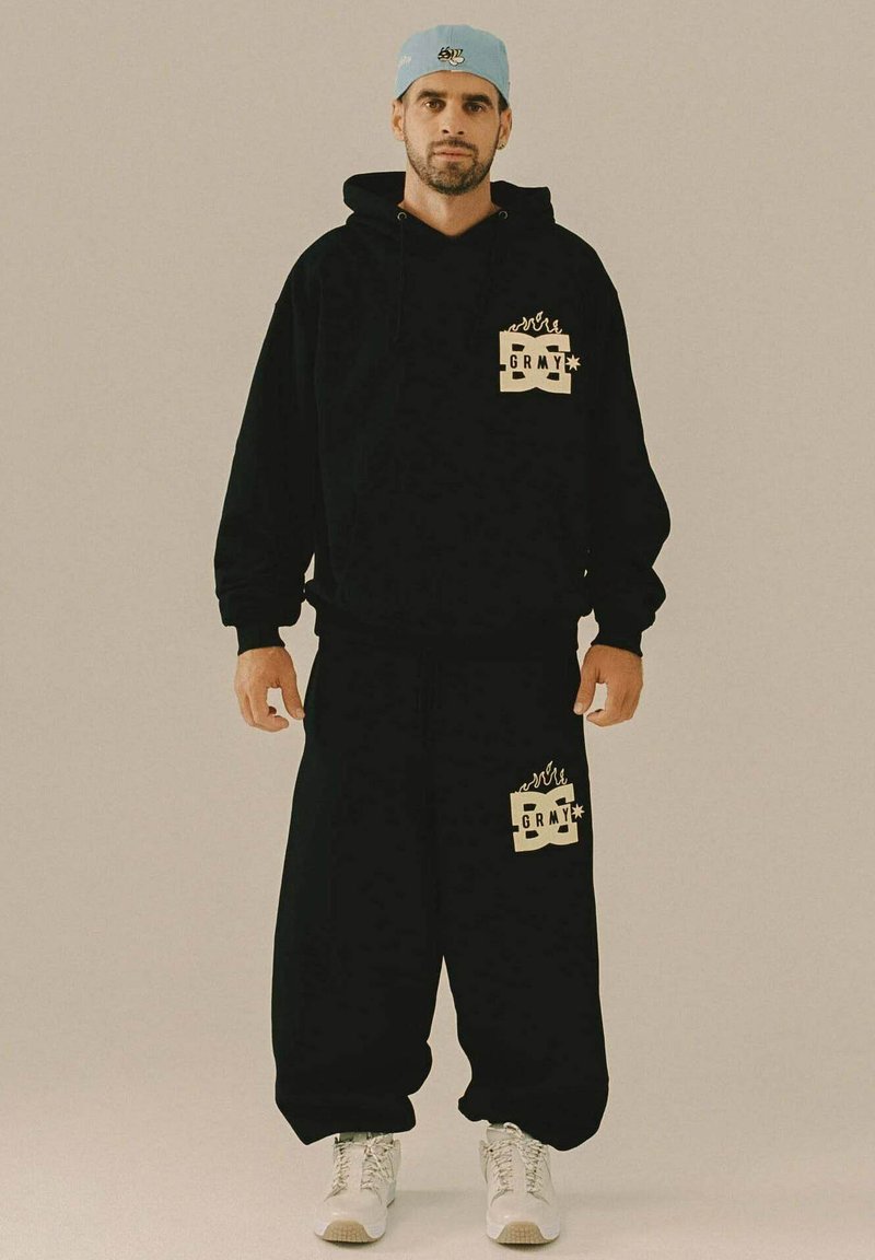 Hombre con sudadera negra y pantalones anchos negros a juego con logotipos, zapatillas blancas y gorra ligera azul al revés, de pie frente a un fondo uniforme.