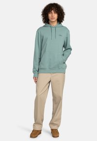 Sudadera verde clara con bolsillo frontal y capucha con cordón, combinada con pantalones beige y mocasines marrón claro. El modelo está de pie contra un fondo blanco.