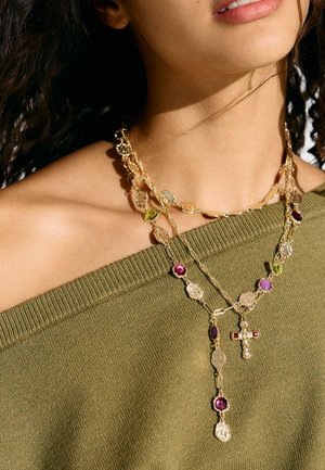 Nainen, jolla on kiharat hiukset, pukeutunut oliivinvihreään olkapäät paljastavaan paitaan ja kerroksellisiin kultaisiin kaulakoruihin, joissa on värikkäitä jalokiviä ja ristinmuotoinen riipus.