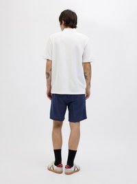 Witte polo met korte mouwen, gestructureerde stof, marineblauwe short, zwarte sokken en casual schoenen met groene accenten en een rood logo.