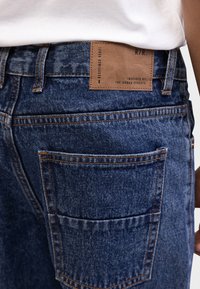 Τζιν παντελόνια σε σκούρο μπλε με υφασμάτινη υφή. Διαθέτουν καφέ δερμάτινο στολίδι με την επιγραφή «REDEFINED REBEL» και δύο πίσω τσέπες με ραφές.