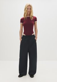 Maroenen bodysuit met korte mouwen en witte bies, gecombineerd met losse, zwarte wijde broek. Model staat met handen in de zakken.