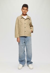 Beige jasje met twee voorzakken en zwarte knopen, gedragen over een wit shirt. Lichtblauwe baggy jeans en witte sneakers maken de outfit compleet.