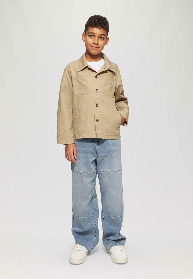 Beige jasje met twee voorzakken en zwarte knopen, gedragen over een wit shirt. Lichtblauwe baggy jeans en witte sneakers maken de outfit compleet.