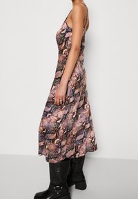 Femme portant une robe sans manches, longue jusqu'aux chevilles, à motif serpentin, associée à des bottes noires montantes, se tenant devant un fond uni.