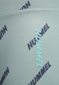 Ljus turkos tyg med en texturerad yta som har ordet "HUMMEL" i kontrasterande mörkblå och aqua tryck i ett diagonalt mönster.