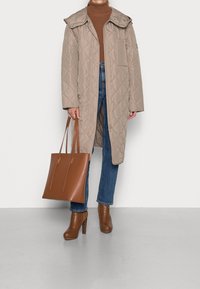 Beiger gesteppter langer Mantel mit großem Kragen, brauner Rollkragenpullover, blaue Jeans, braune Stiefeletten mit hohem Absatz und eine braune Tragetasche.