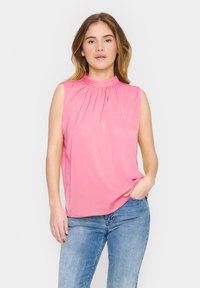Top rosa corto senza maniche con collo alto arricciato, realizzato in tessuto morbido, abbinato a jeans azzurri chiari.