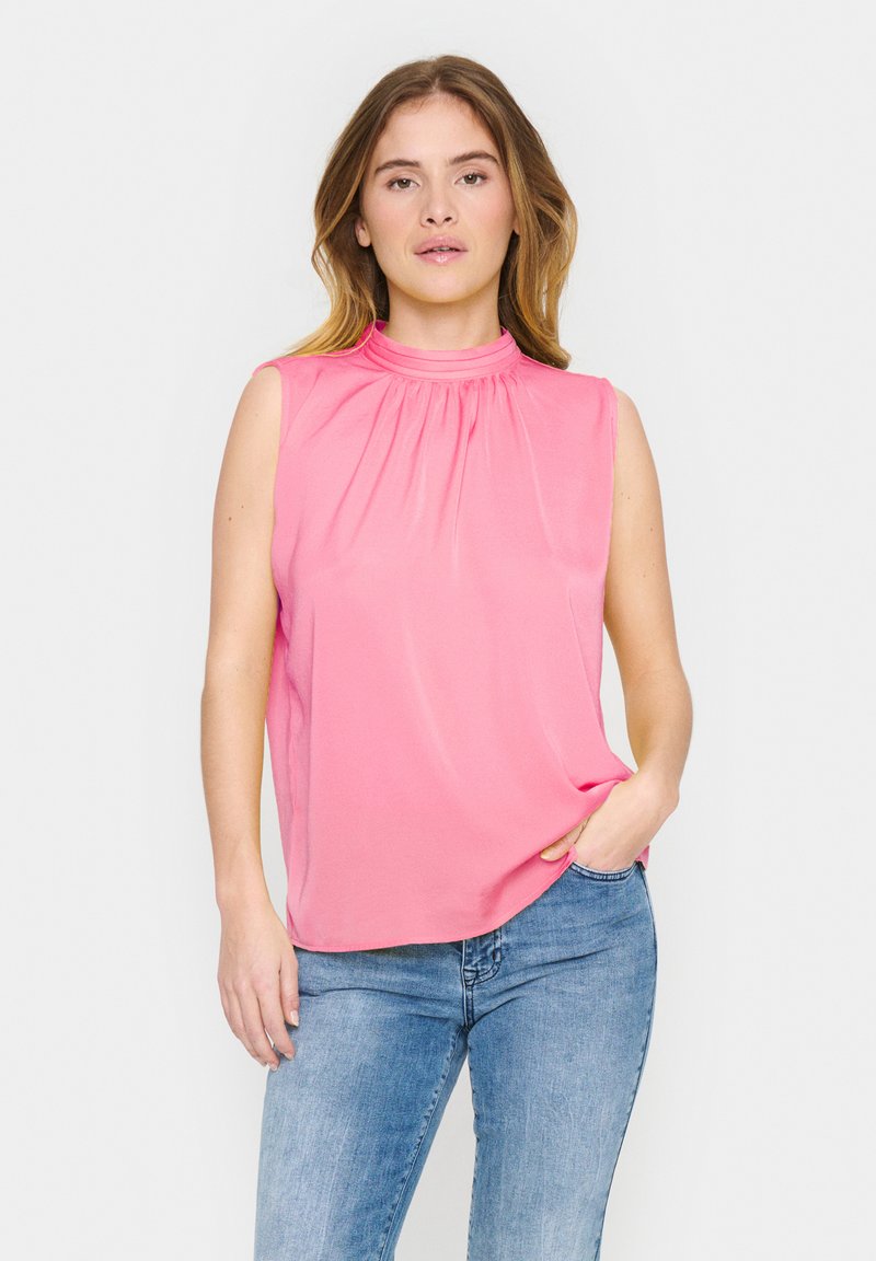 Top rosa corto senza maniche con collo alto arricciato, realizzato in tessuto morbido, abbinato a jeans azzurri chiari.