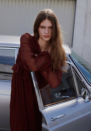 Jeune femme aux longs cheveux portant une robe bordeaux transparente, appuyée sur la porte ouverte d'une voiture vintage argentée, sur un fond de mur neutre.