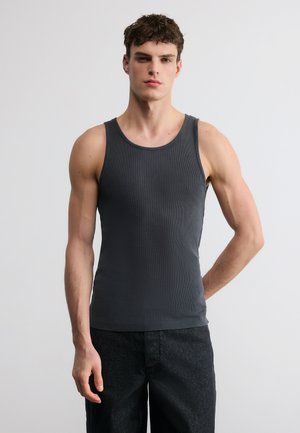 Jeune homme debout, un bras derrière le dos, portant un débardeur gris foncé sans manches et un pantalon noir, sur un fond clair uni.