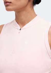 Gros plan d'une personne portant un haut sans manches rose clair avec une courte fermeture éclair à l'avant et un petit logo blanc sur la poitrine.