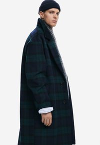 Plaid wool coat in navy and green tones, featuring a notched collar, double-breasted buttons, and side pockets. Textured knit underlayer visible.