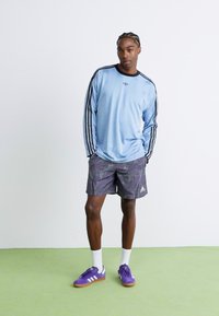 Mies, joka seisoo käsivarret taskuissa, päällään vaaleansininen pitkähihaisen Adidas-paita, kuviolliset sortsit, valkoiset sukat ja violetit Adidas-tennarit.