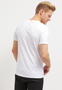 White cotton t-shirt with short sleeves, round neck, and a smooth texture, worn backward, showing a clean silhouette and no visible graphics.