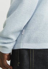 Suéter de textura azul claro con un dobladillo acanalado. Se observa una etiqueta de la marca visible en la costura lateral. Jeans de mezclilla oscuros con costuras doradas se ven abajo.