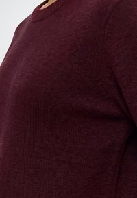 Gros plan d'une personne portant un pull tricoté uni de couleur rouge foncé avec un col rond sur un fond clair.