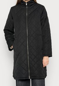 Svart quiltad lång jacka med framdragkedja, stående hållning, avslappnade händer, parat med mörka jeans.