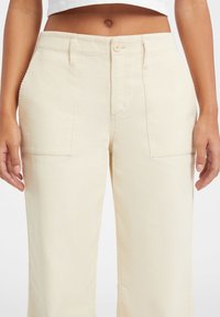 Parte inferiore del corpo che indossa pantaloni in corduroy crema a vita alta con tasche frontali a toppa, chiusura a bottone e un top bianco corto.