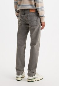 Personne portant un jean droit gris, une chemise à rayures beige et bleues à manches longues et des baskets blanches avec des accents verts.