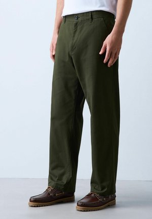 Mann trägt olivgrüne gerade geschnittene Hose, braune Leder-Bootsschuhe und ein weißes Hemd, steht auf einem hellgrauen Boden.