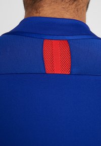 Sininen urheilupaita, jossa on teksturoitu punainen verkkopaneeli takakolmannessa kauluksessa. Kangas on sileä ja istuva, klassisella muotoilulla.