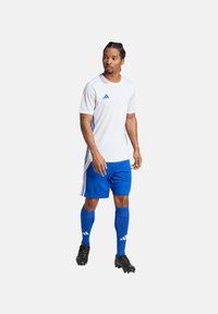Camiseta atlética blanca con detalles azules, combinada con pantalones cortos azules, calcetas azules hasta la rodilla y botas negras. Detalle de tres rayas en los lados.