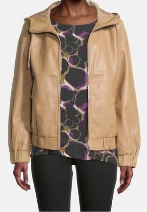 Frau trägt eine beige Reißverschlussjacke über einem mehrfarbigen, abstrakt gemusterten Hemd und schwarzen Hosen, gezeigt vom Hals bis zur Hüfte.