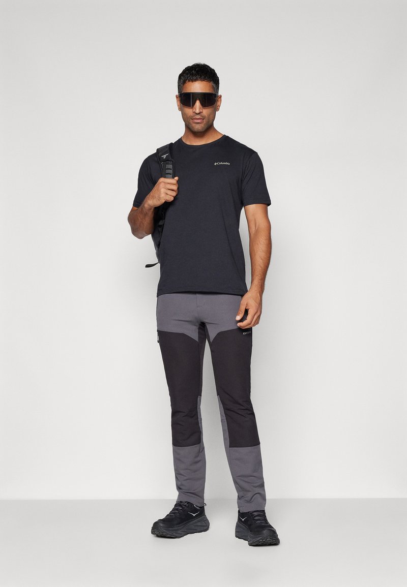 Camisa negra de manga corta, pantalones grises y negros con paneles en las rodillas, zapatillas negras y una mochila. Tejido suave y corte atlético.