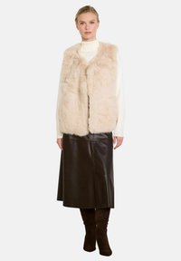 Gilet in pelliccia sintetica di un beige chiaro con una texture morbida, indossato sopra un dolcevita crema, abbinato a una gonna in pelle marrone e stivali alti marroni fino alla coscia.