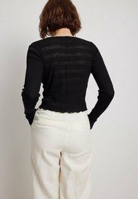 Schwarzes, geripptes Langarmoberteil mit kurzem Schnitt und gewelltem Saum. Hat horizontale Streifen und eine körpernahe Silhouette. Wird mit weißen Hosen getragen.