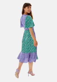 Floral print dress with short flutter sleeves, green and purple patterns, cinched waistline, and a ruffled hem, paired with light purple heels.