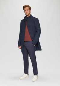 Marineblauwe wollen jas met een hoge kraag, knoopsluiting, gecombineerd met een bordeauxrode trui en marineblauwe broek. Witte sneakers maken de outfit af.