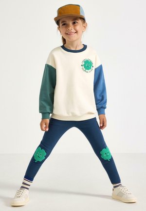 Barn iført en cremefarvet sweatshirt med grønne og blå ærmer og marineblå bukser med smilende grønne kløverpletter på knæene, stående med benene fra hinanden.