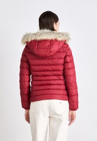 Červená puffa bunda s kožúškom pod kapucňou, prešívaný dizajn a rebrované manžety. Kombinovaná s béžovými nohavicami, ktoré ukazujú priliehavý strih.