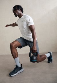 Sportsudøver i en hvid tætsiddende skjorte og grå shorts udfører et udstræk med en sort kettlebell. Grå sportssko og hvide sokker.