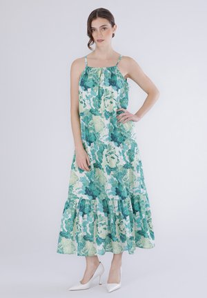 Femme debout portant une robe mi-longue sans manches à fleurs vertes et blanches avec de fines bretelles et des chaussures blanches à talons hauts, sur un fond uni.