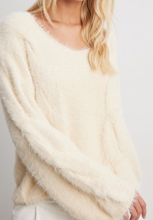 Personne blonde portant un pull doux, duveteux de couleur crème avec des manches longues, visible des épaules aux hanches sur un fond uni.