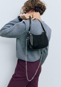 Borsa a tracolla in pelle nera con forma arrotondata, tracolla in catena argentata e superficie liscia e lucida. Indossata con un maglione grigio e pantaloni bordeaux.