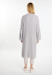 Cardigan lungo e textured in grigio chiaro, con una vestibilità ampia e maniche larghe. Indossato sopra pantaloni corti chiari e scarpe slip-on.
